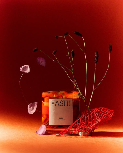 Bougie Aura Yashi sur fond rouge, entre tiges séchées, éclats roses et sculpture en résille rouge