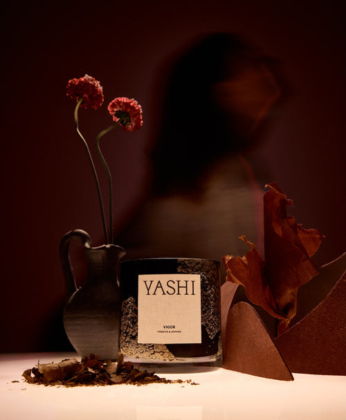 Bougie Vigor Yashi en lumière chaude, près d'un pichet noir, de fleurs séchées et de feuilles brunes
